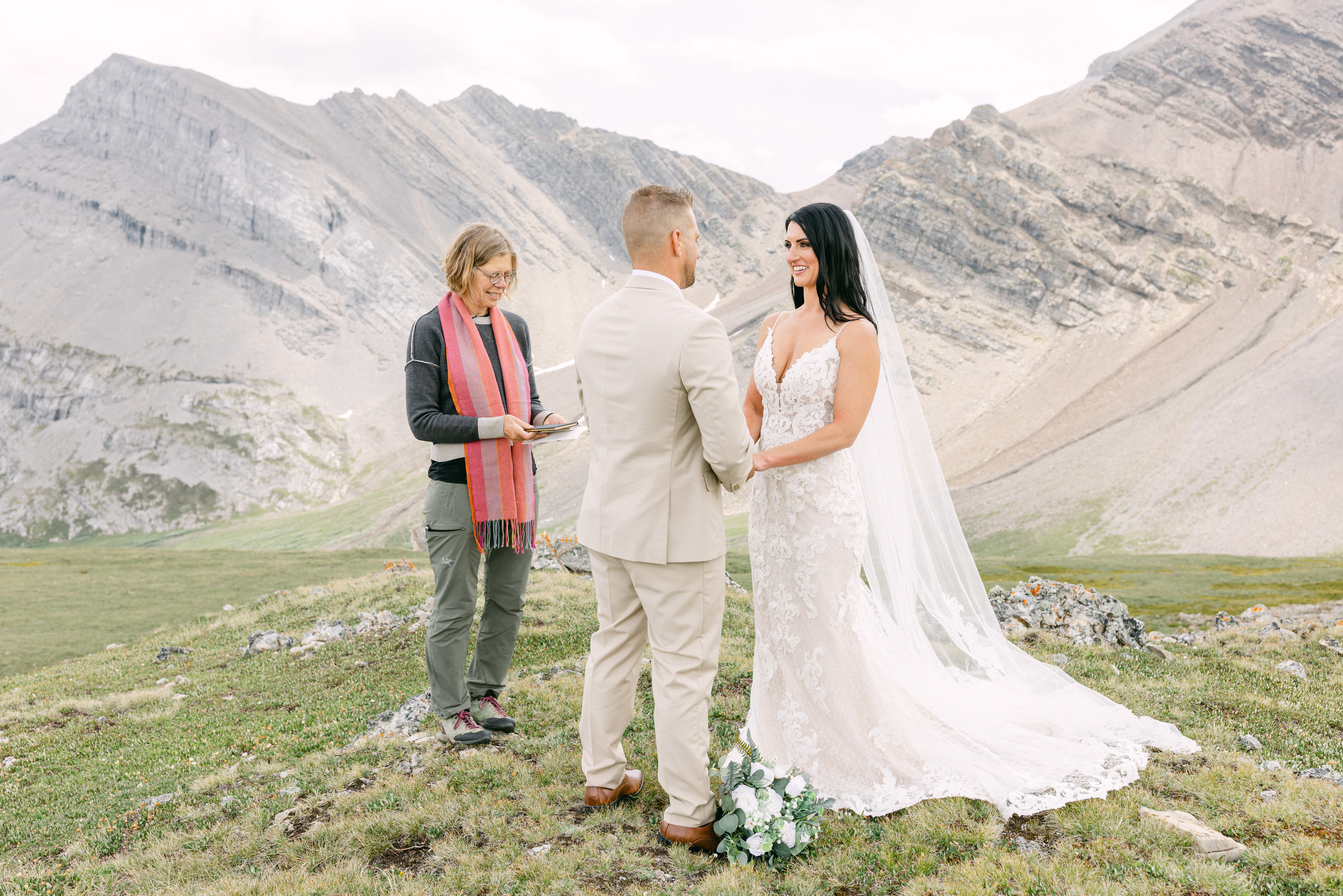 A bride and groom exchange vows in a scenic outdoor setting surrounded by mountains, with an officiant holding a book.