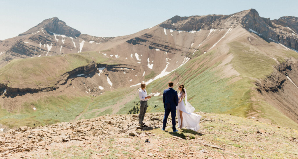 A couple exchanges vows during an outdoor wedding ceremony on a scenic mountain peak, with dramatic mountain landscapes in the background.