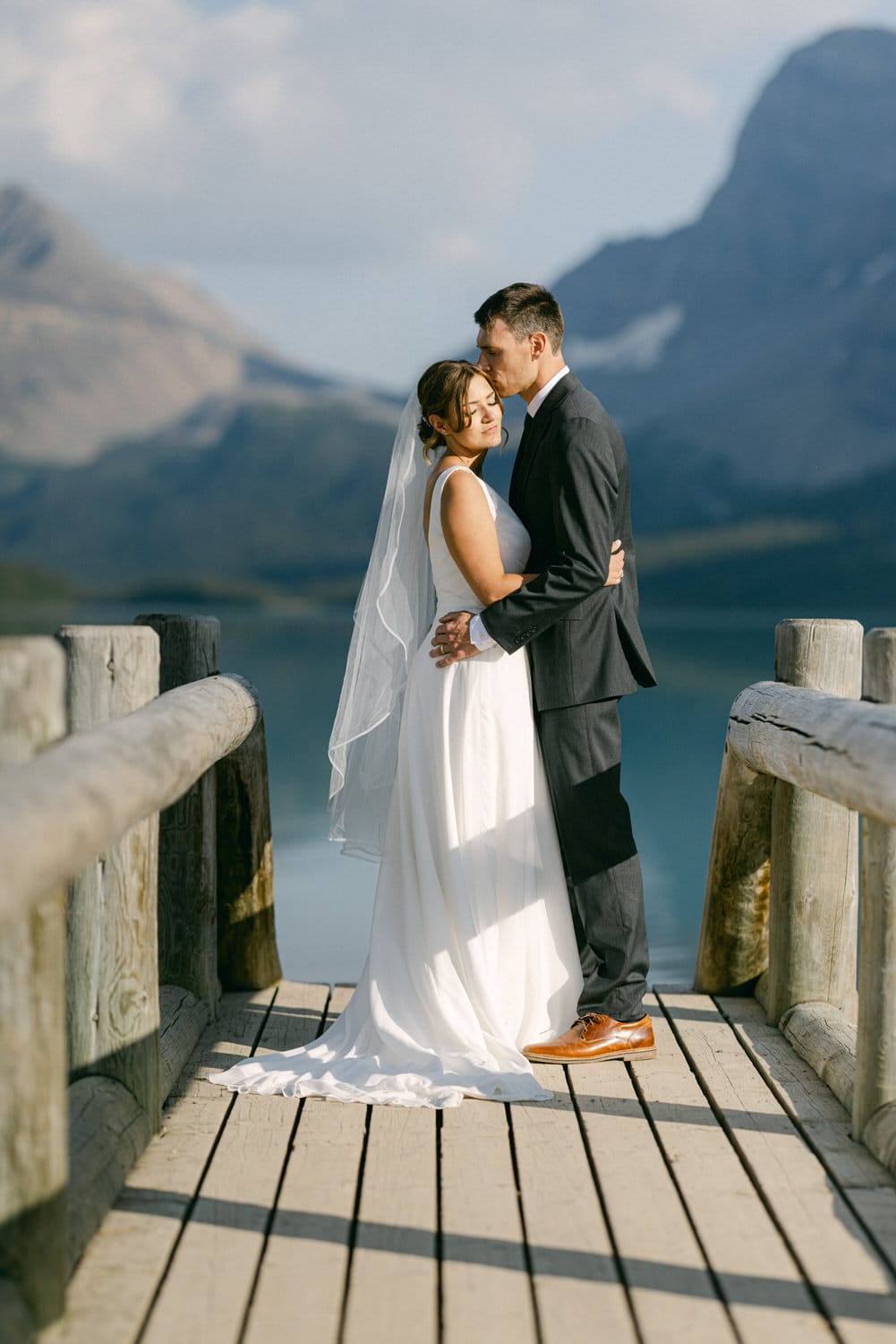 A couple embraces on a wooden dock overlooking a serene lake and mountains, capturing a tender moment on their wedding day.