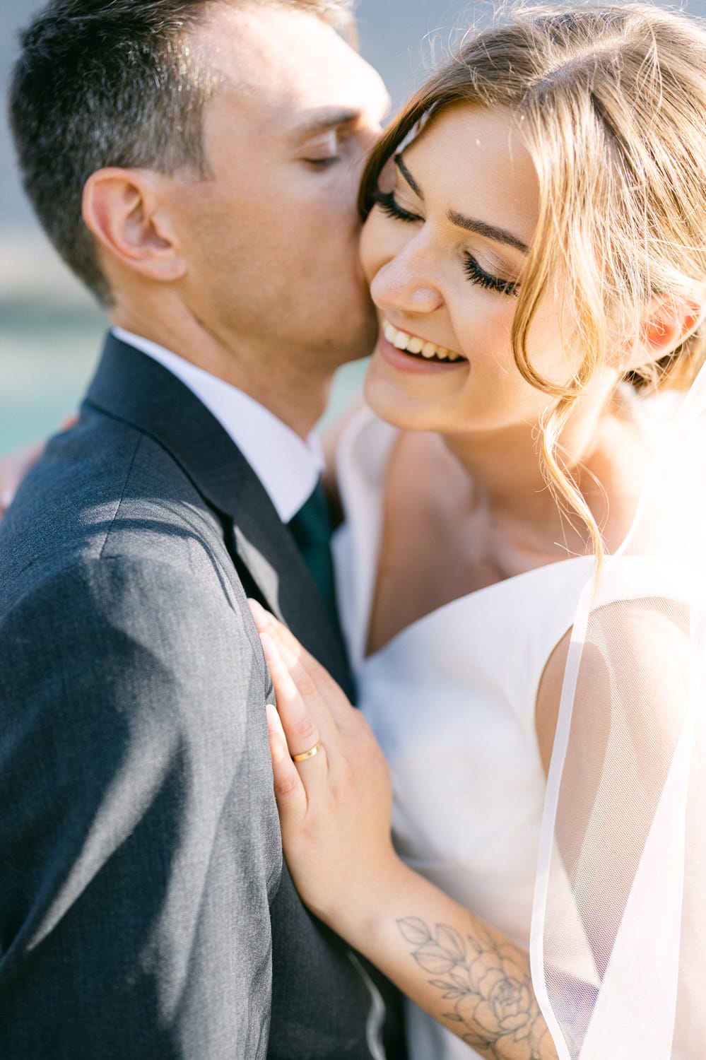 A joyful couple sharing a loving embrace, with the groom kissing the bride's cheek as she smiles radiantly, showcasing her wedding attire and delicate tattoo.