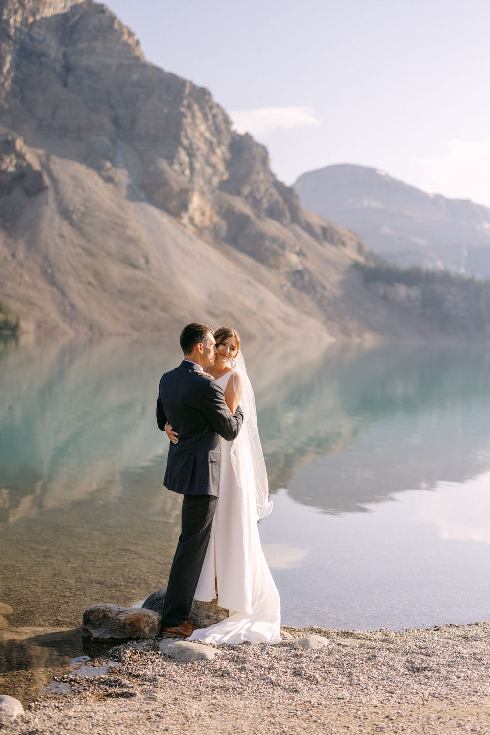 A couple embraces by a tranquil lake surrounded by mountains, capturing a moment of love and serenity.