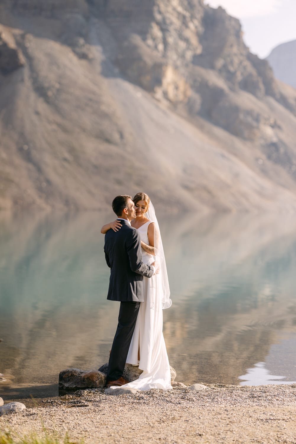 A couple embraces on a rocky shore beside a serene lake, surrounded by mountains, capturing a romantic wedding moment.