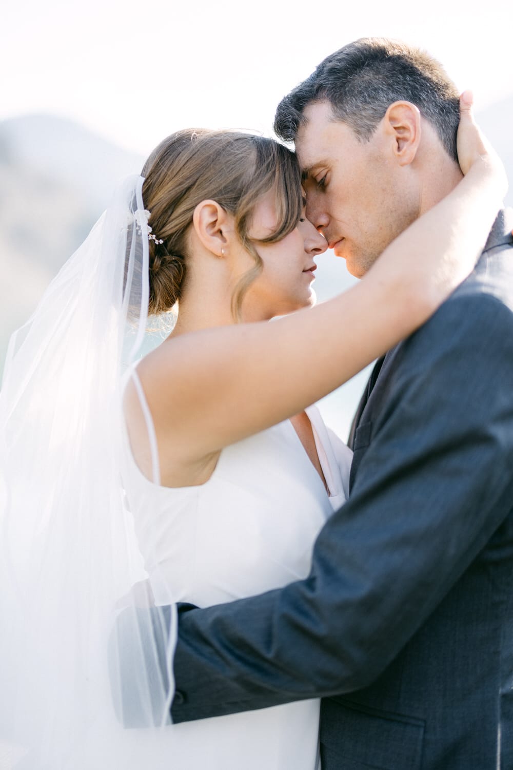 A bride and groom share a tender moment, forehead to forehead, surrounded by soft natural light.