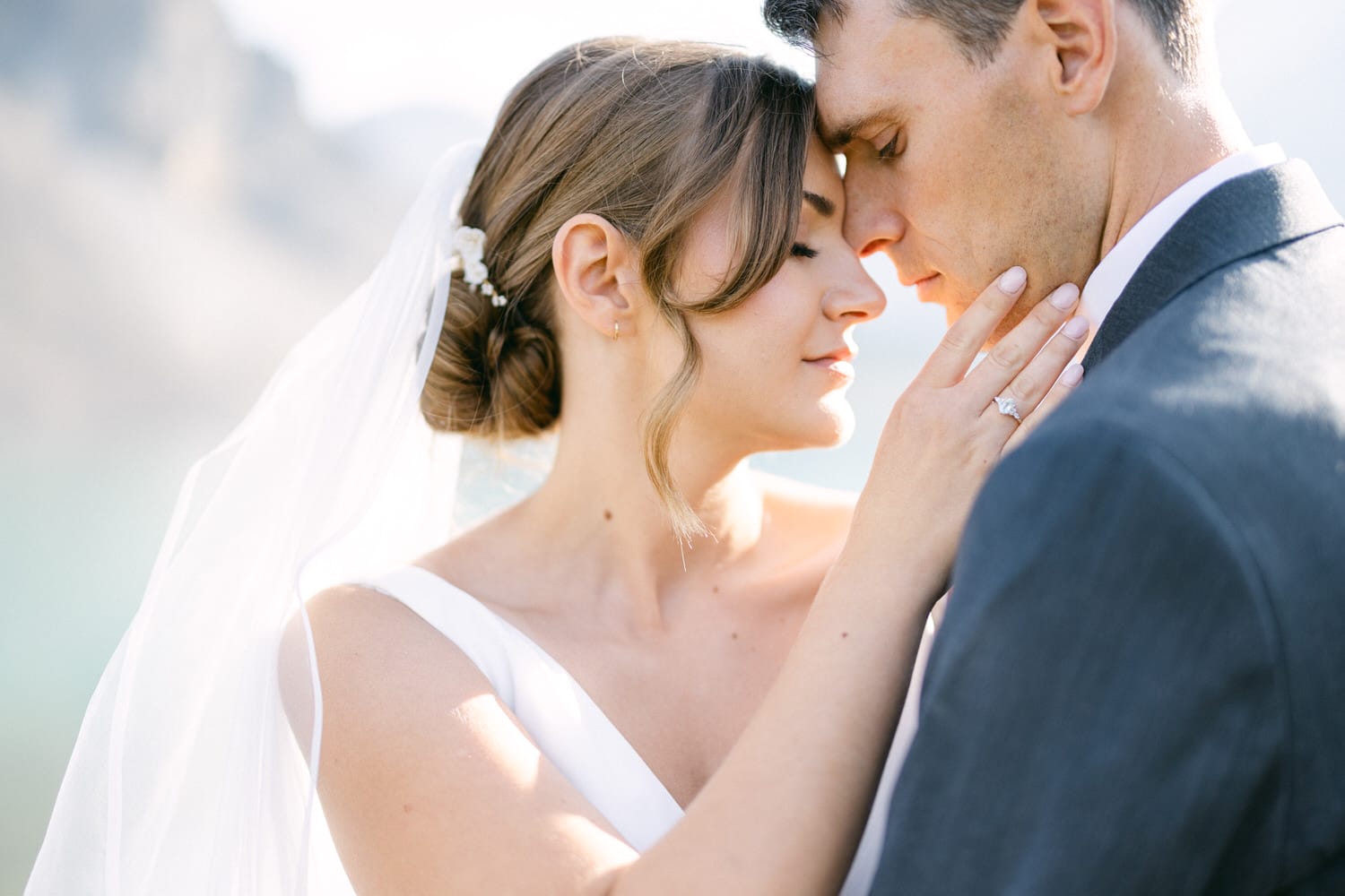 A close-up of a bride and groom sharing an intimate moment, with the bride's hand touching the groom's face, both gazing at each other against a soft, blurred background.