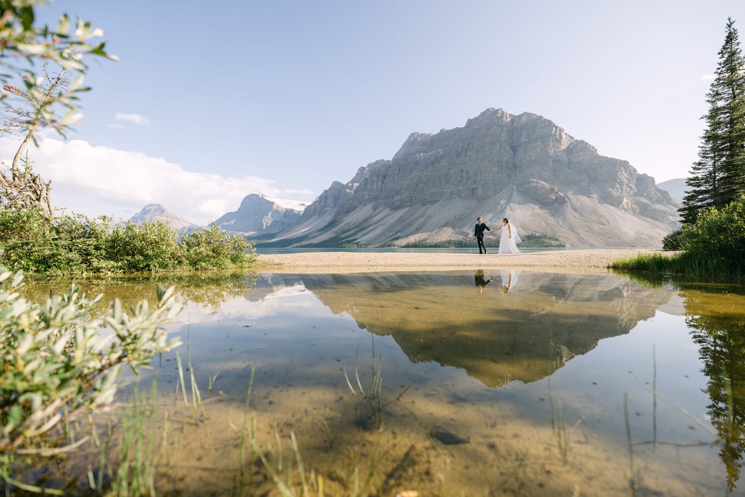 A couple stands hand-in-hand by a serene lake, with majestic mountains reflected in the water under a clear sky.