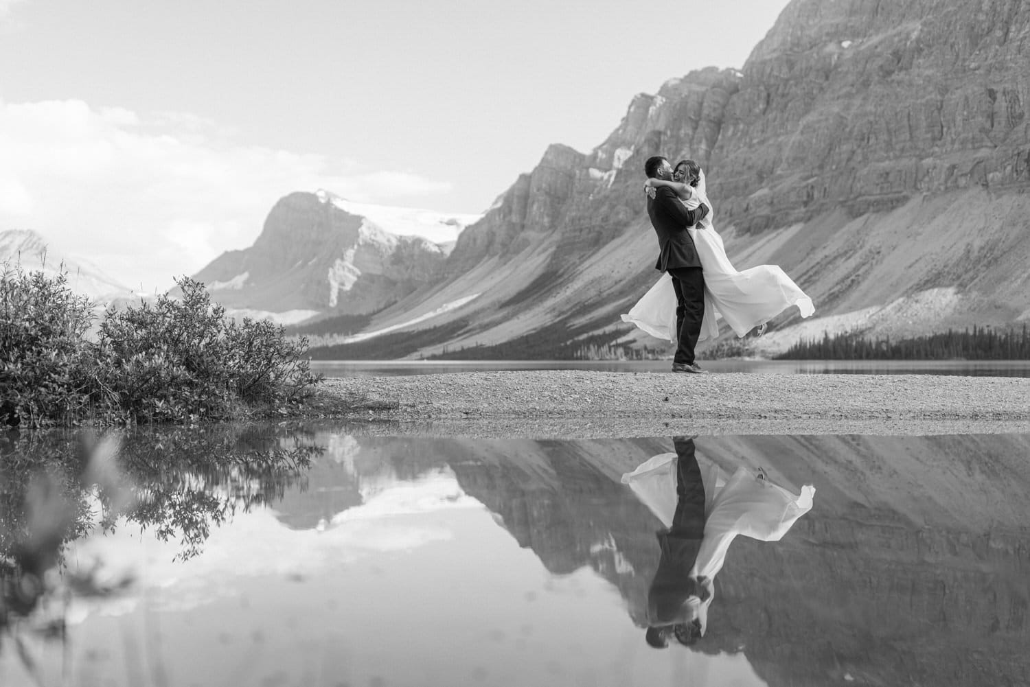 A couple embraces joyfully in a stunning mountainous landscape, reflecting in a nearby water puddle.