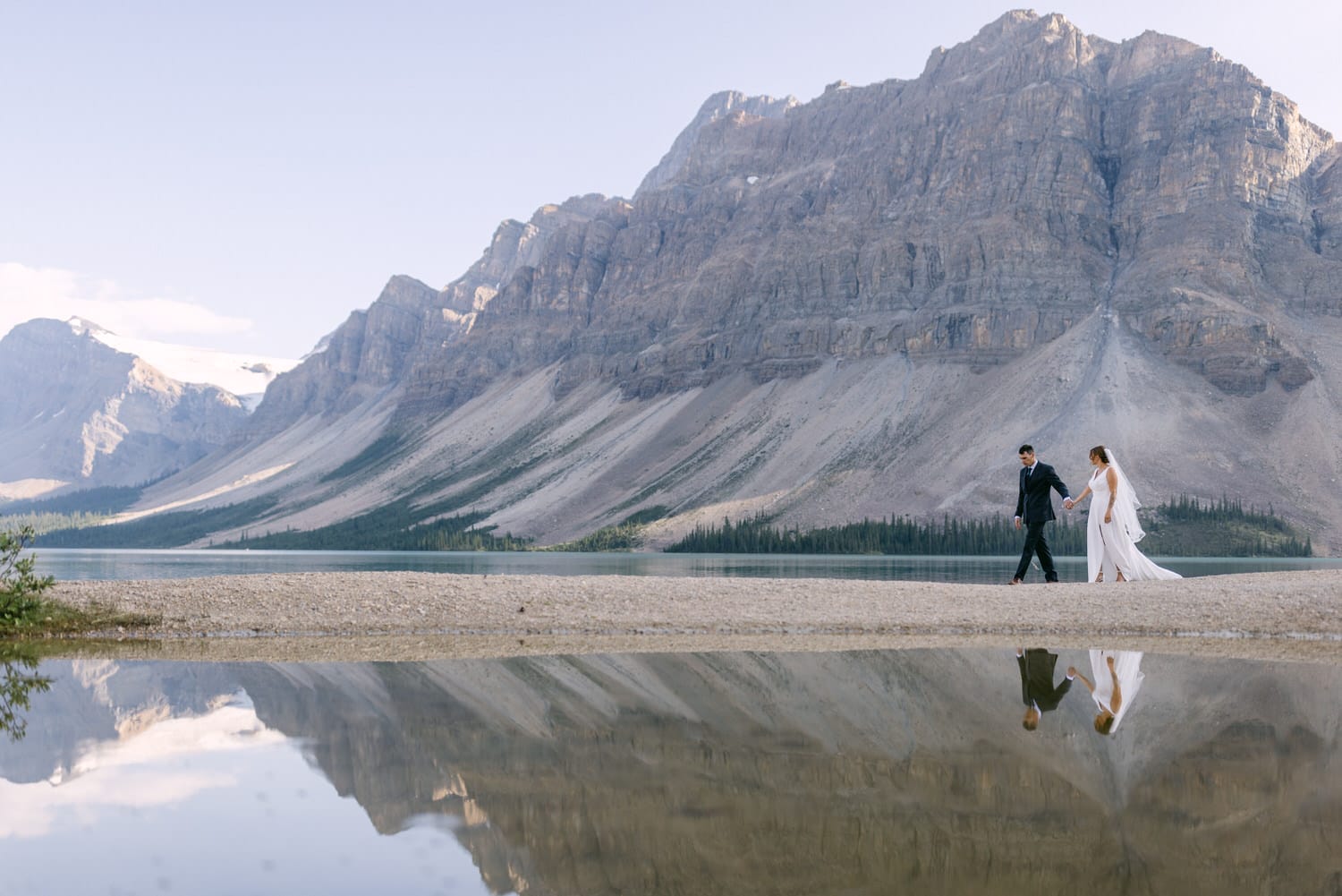 A couple walks hand in hand along a sandy shore with mountains reflected in the calm water.