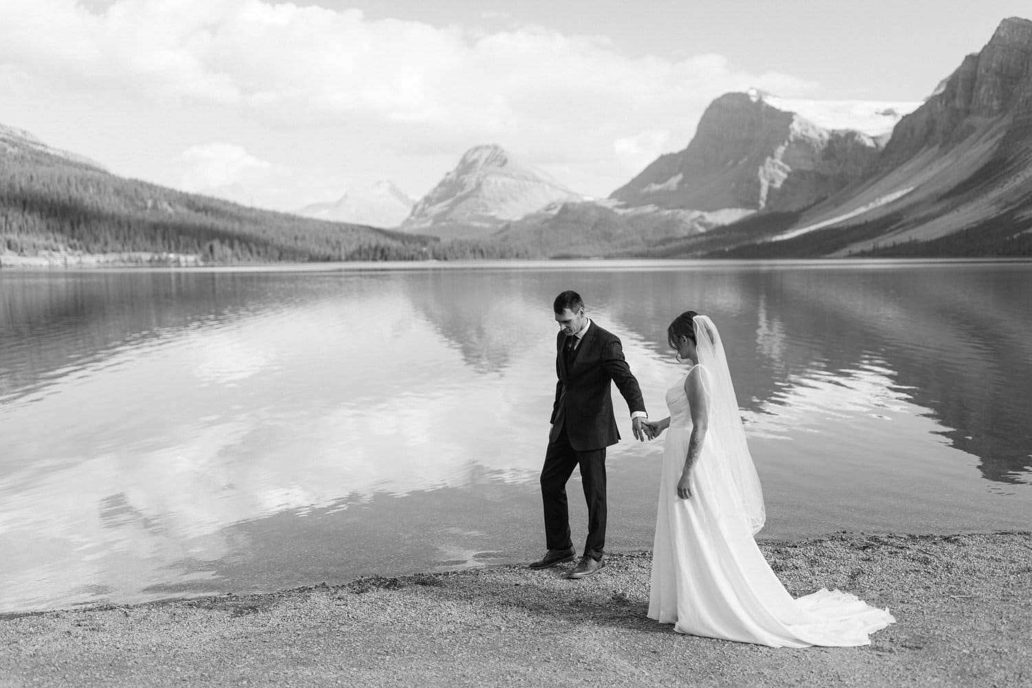 A couple, dressed in wedding attire, holds hands while walking along a serene lake shore, surrounded by stunning mountains and a clear sky.