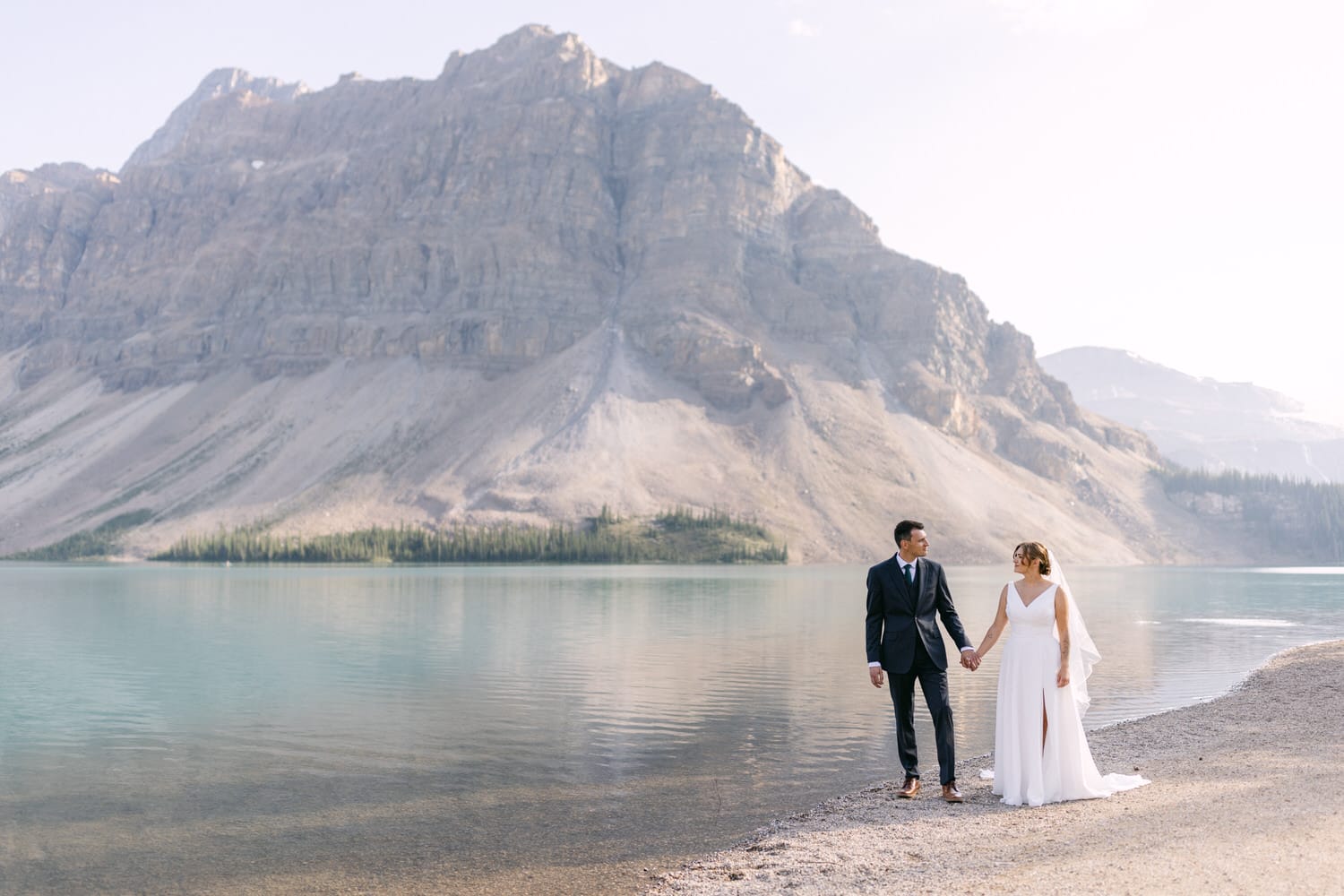 A bride and groom hold hands by a serene mountain lake, with towering cliffs and lush greenery in the background.