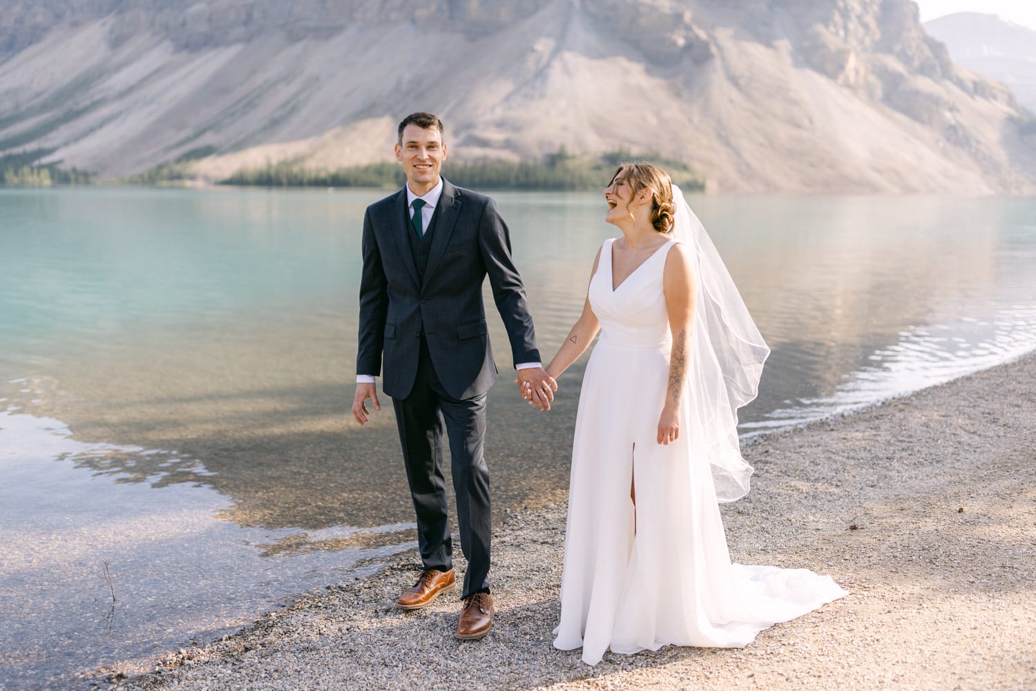 A joyful bride and groom holding hands by a serene lake, with mountains in the background.