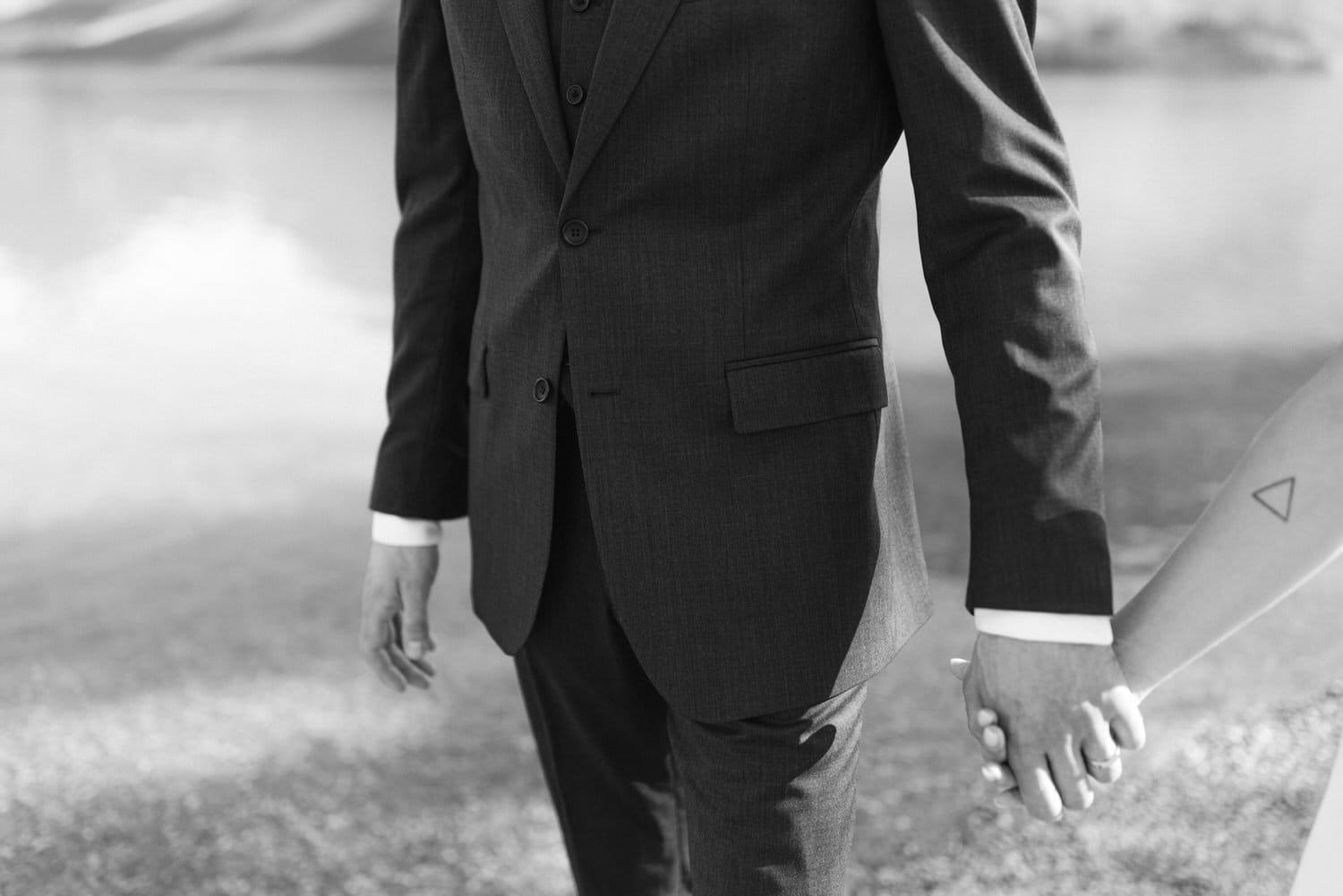 A close-up of a man in a formal suit holding hands with a woman, showcasing their connection against a serene outdoor backdrop.