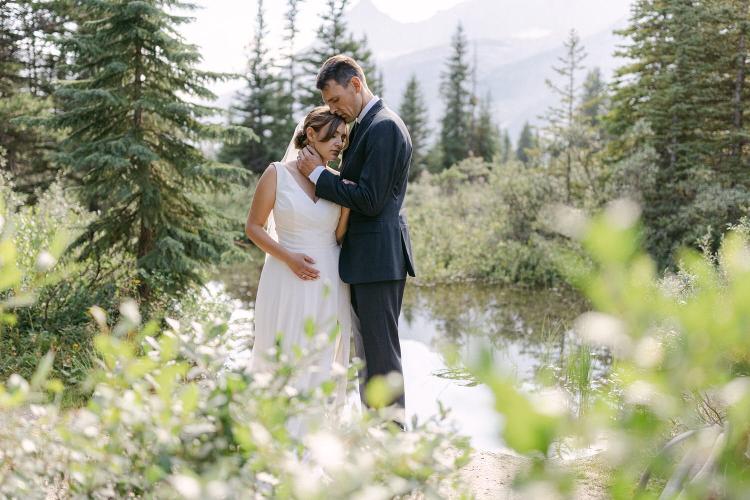 A couple stands close together in a serene natural setting, surrounded by trees and a calm water body, displaying a moment of intimacy.