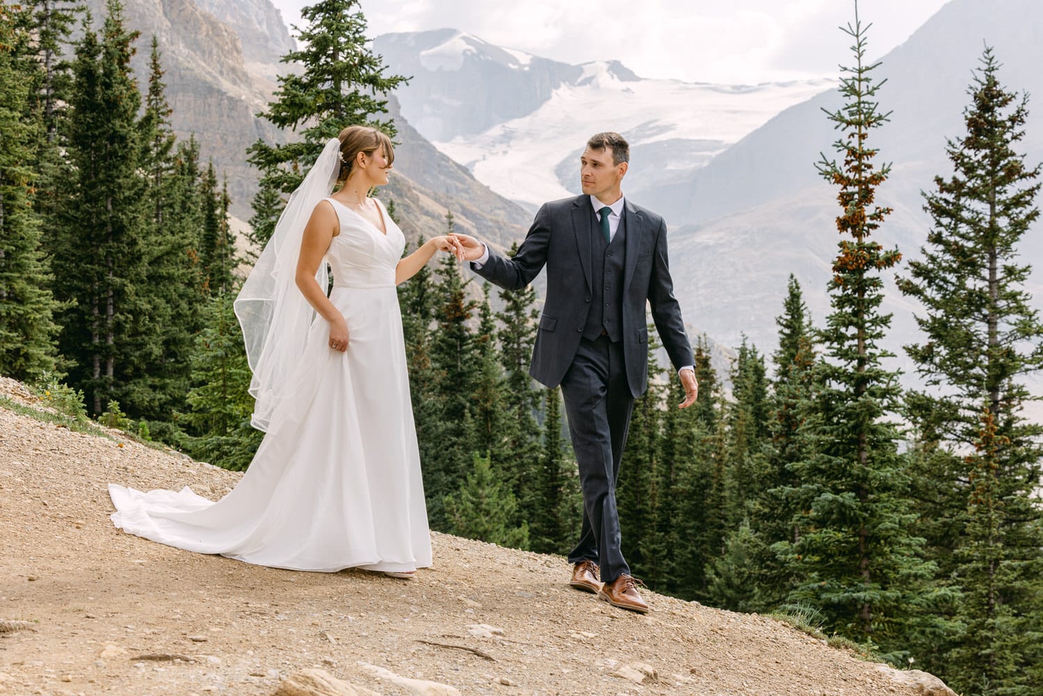 A bride and groom walk hand in hand along a scenic mountain path, surrounded by lush evergreen trees and a distant glacier.