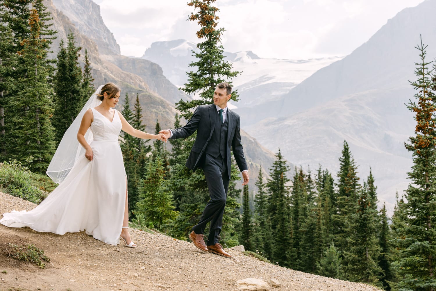 A couple walks hand in hand along a scenic trail, surrounded by lush trees and majestic mountain landscapes during their wedding day.