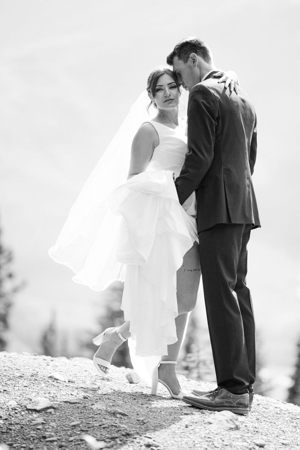 A bride and groom pose intimately, with the bride wearing a flowing white dress and veil, while the groom leans in closely, set against a nature backdrop.