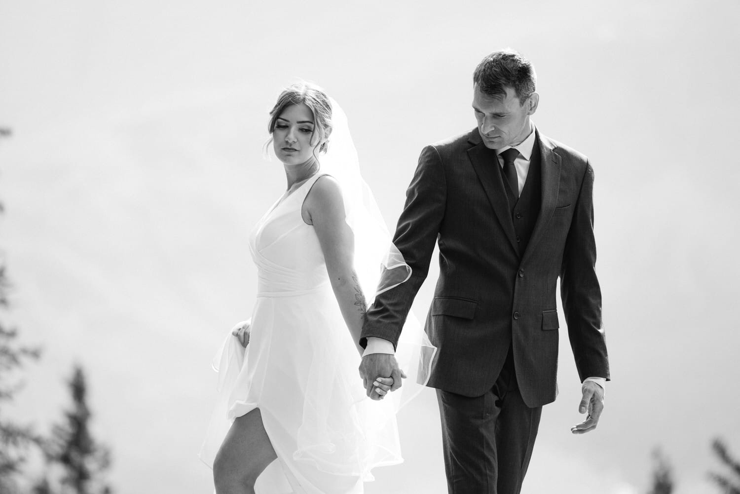 A couple holding hands during an intimate moment, the bride in a flowing white dress and veil, while the groom wears a dark suit, set against a soft, blurred backdrop.