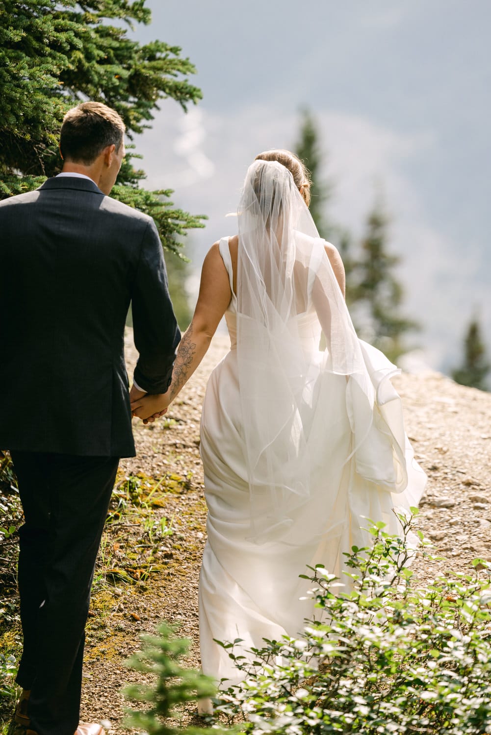 A bride and groom walk hand in hand through a scenic outdoor path, surrounded by trees and a soft natural landscape.