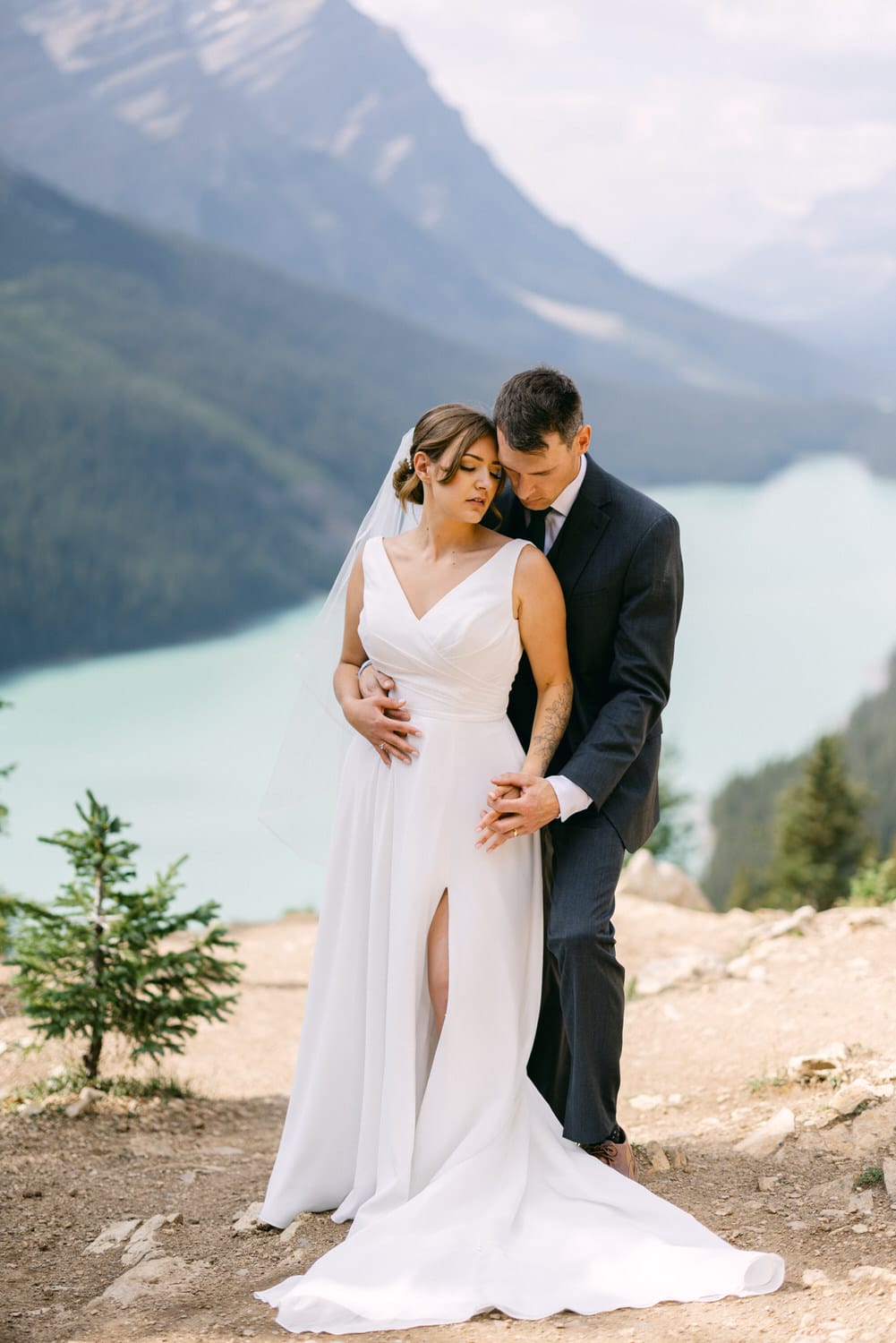 A couple embraces in a stunning mountain landscape, with a serene lake in the background, showcasing the bride's elegant gown and the groom's tailored suit.