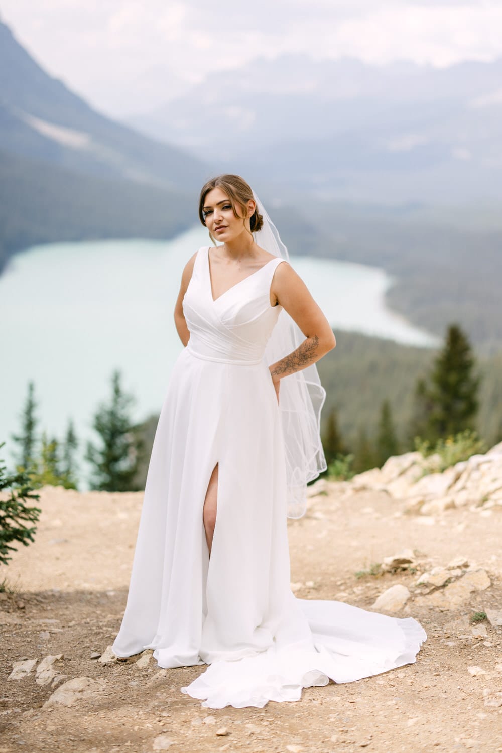 A bride in a white dress poses confidently on a rocky trail with a stunning lake and mountains in the background.