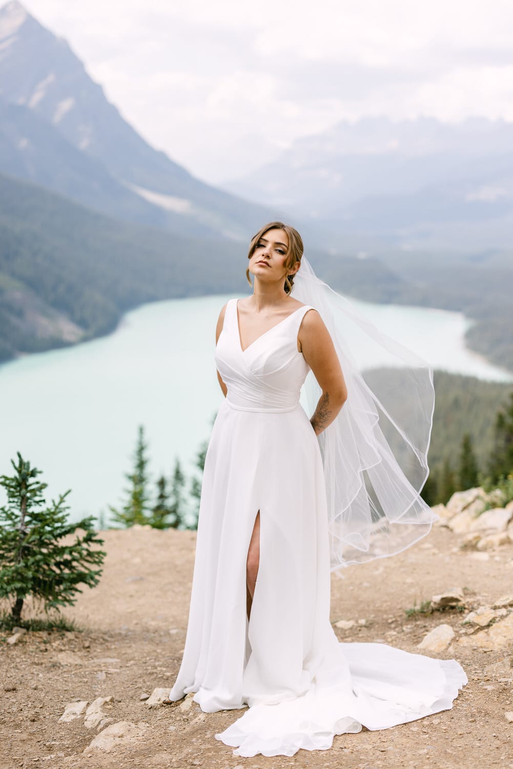 A bride in a flowy white dress and veil poses confidently against a breathtaking mountain lake backdrop, showcasing a blend of nature and elegance.