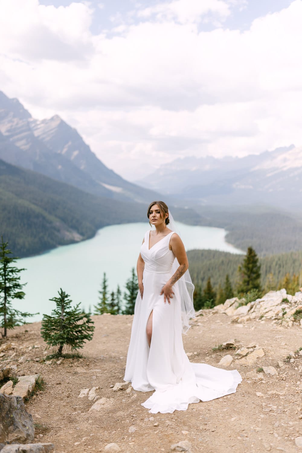 A bride in a flowing white gown poses elegantly against a breathtaking mountain backdrop with a turquoise lake below.