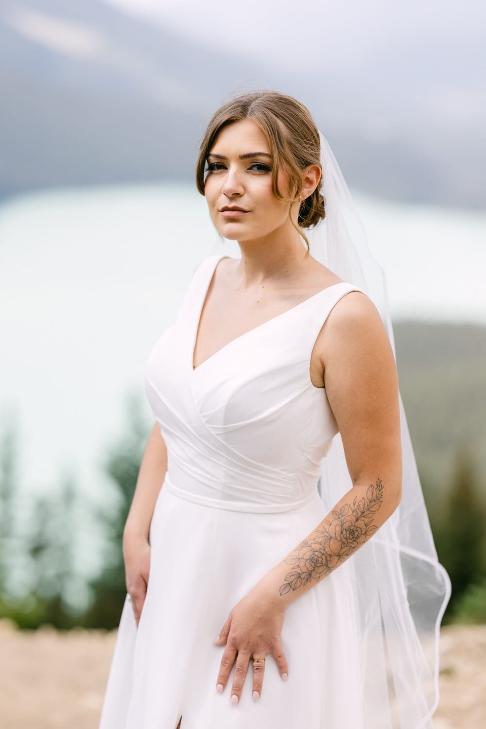 A bride in a white dress with a veil poses confidently in front of a beautiful mountainous backdrop, showcasing her tattoo and ring.