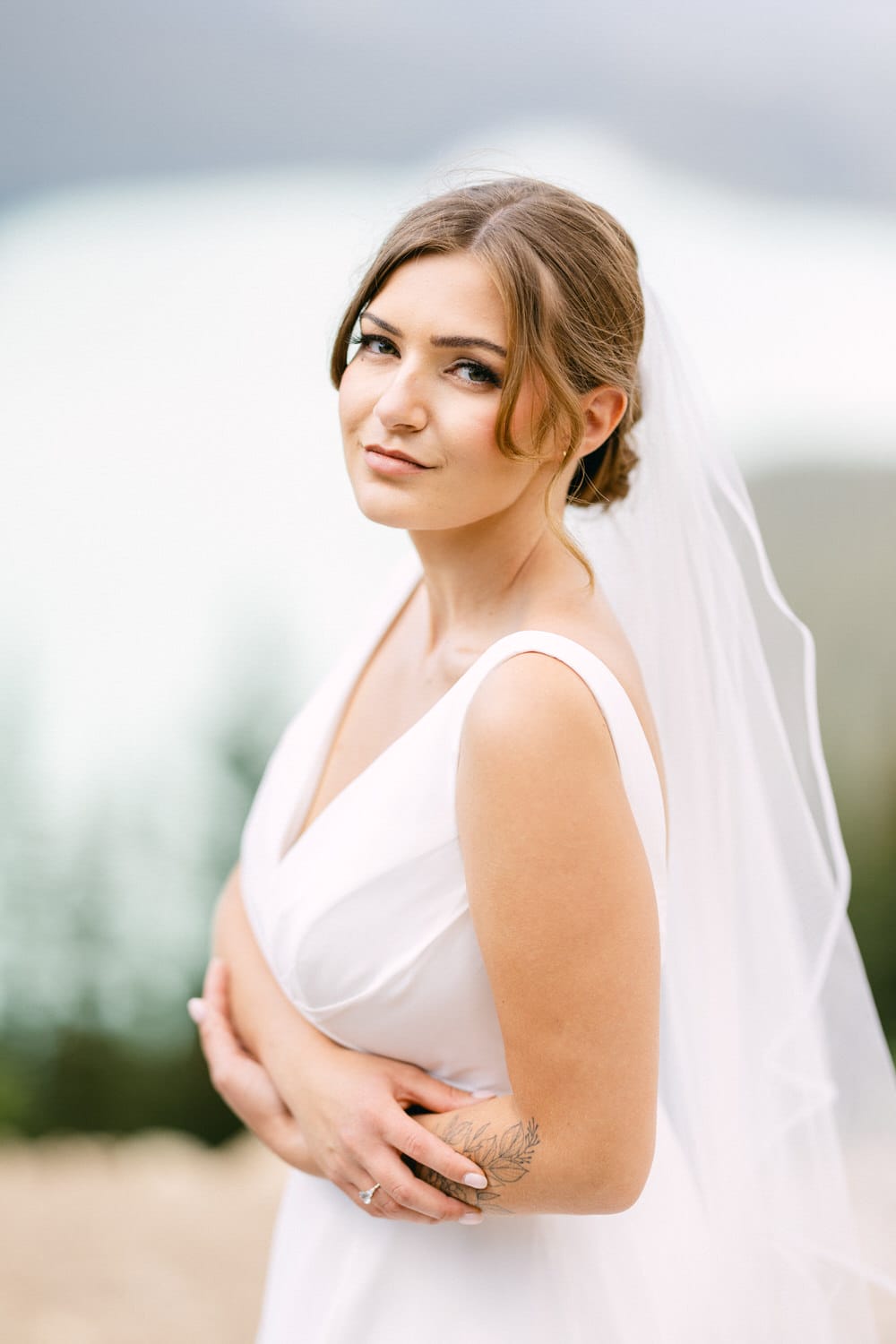 A beautiful bride in a simple white gown and veil poses confidently, showcasing her arm tattoo against a backdrop of soft blue and green hues.