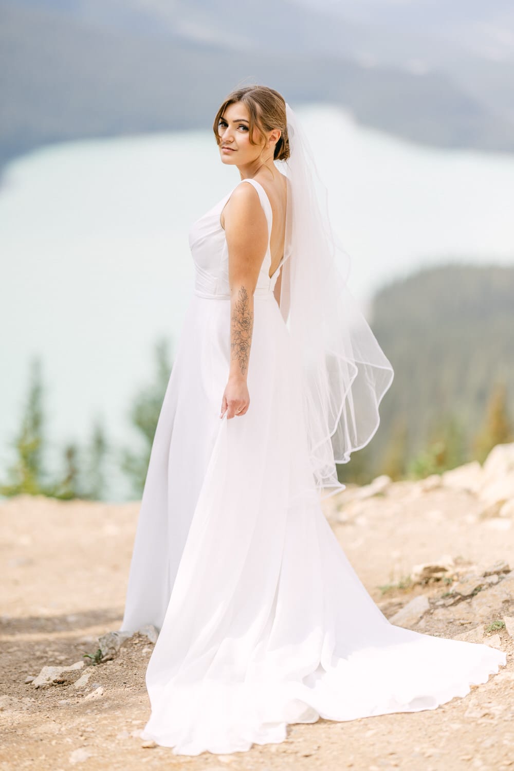 A bride in a flowing white gown and veil stands gracefully against a scenic backdrop of mountains and a serene lake.