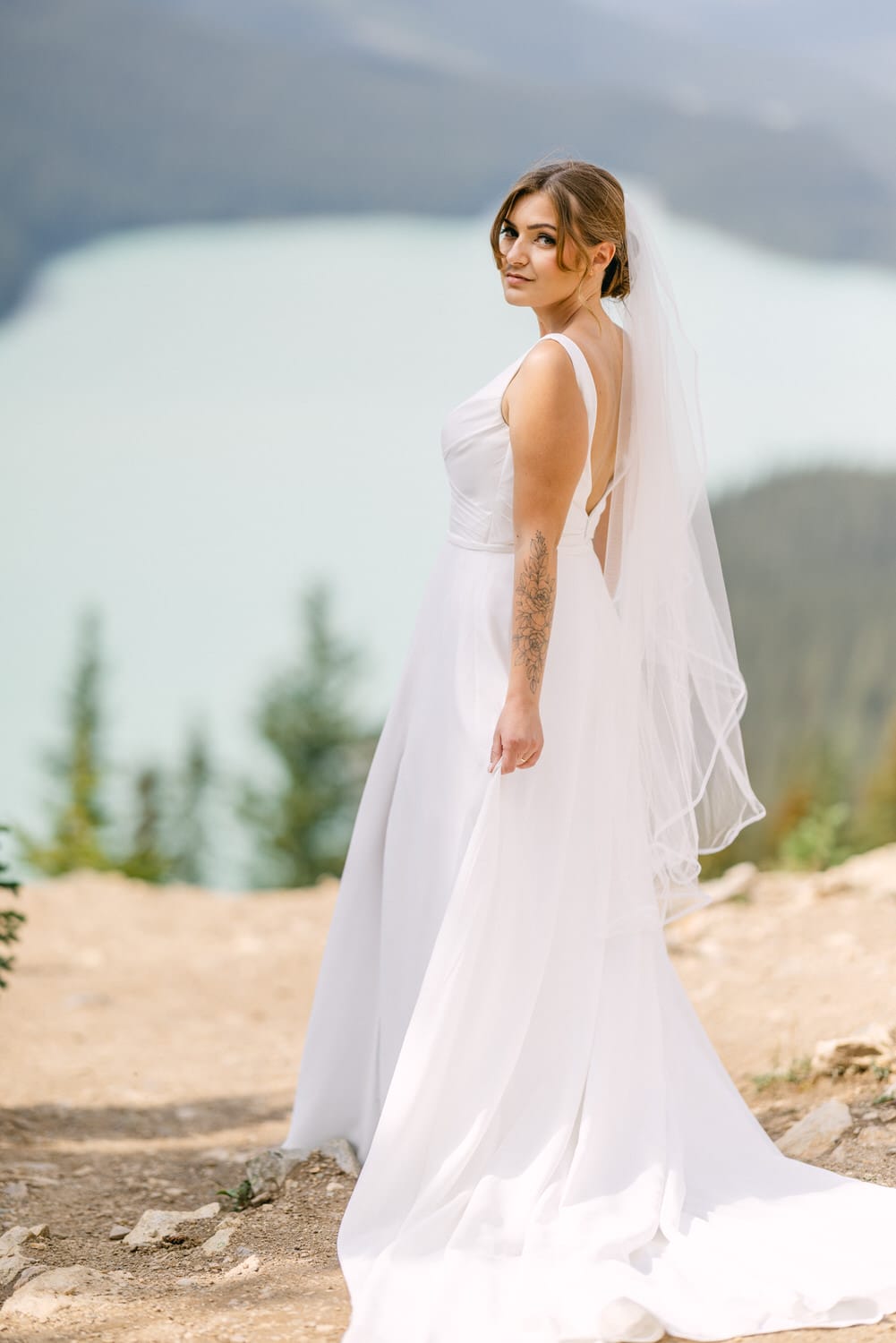 A bride in a flowing white gown with a veil stands gracefully on a rocky outcrop, overlooking a serene lake and lush greenery in the background.