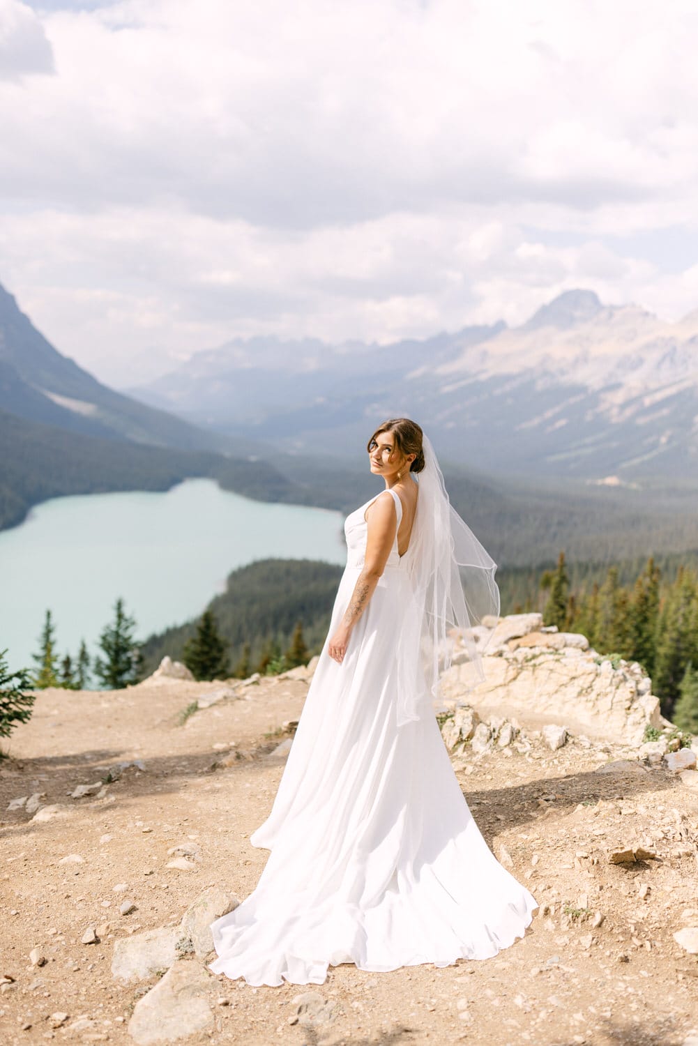 A bride in a flowing white dress and veil stands on a rocky ledge, overlooking a serene turquoise lake surrounded by mountains and trees.