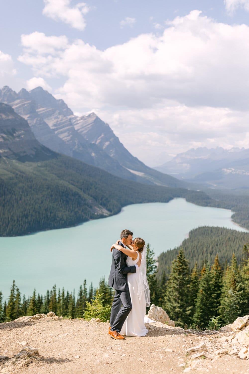 A couple shares a kiss during their wedding ceremony, surrounded by stunning mountains and a turquoise lake.