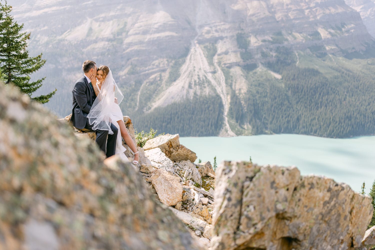 A couple shares a tender moment on a rocky ledge with a stunning mountainous landscape and turquoise lake in the background.