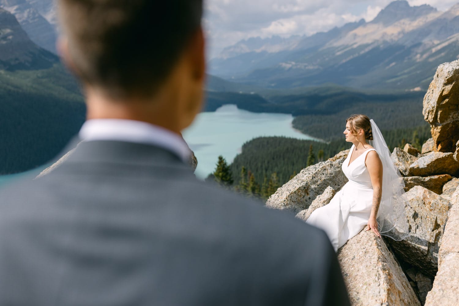 A bride in a white dress and veil sits on rocky terrain, gazing out at a stunning mountainous landscape while a groom is seen from behind, slightly blurred.