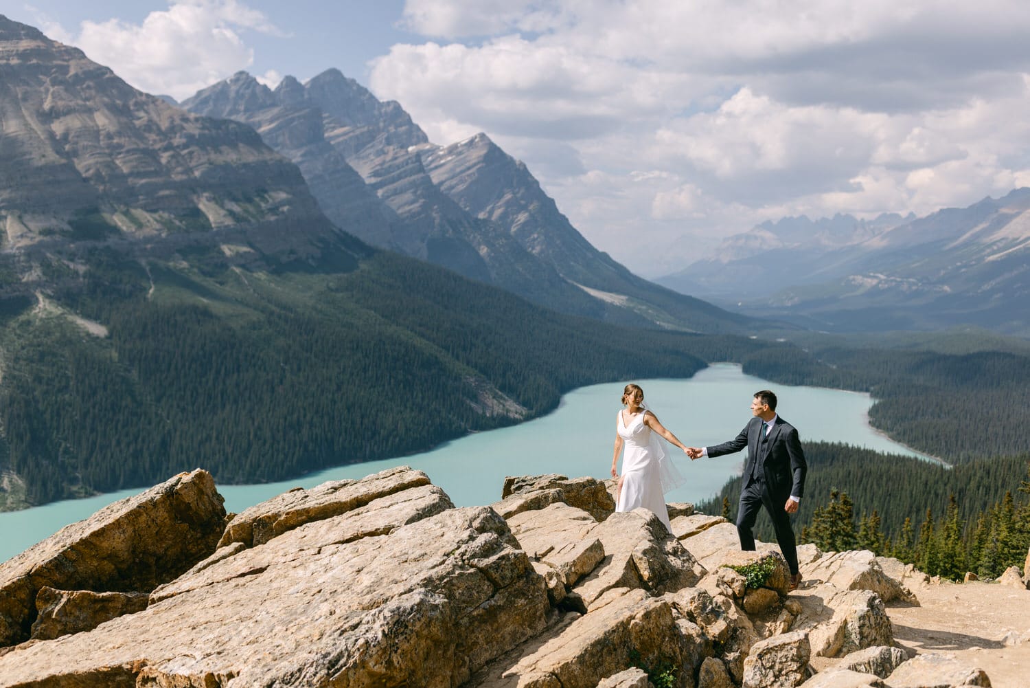 A couple holds hands while standing on rocky terrain overlooking a serene lake and rugged mountains in the background.