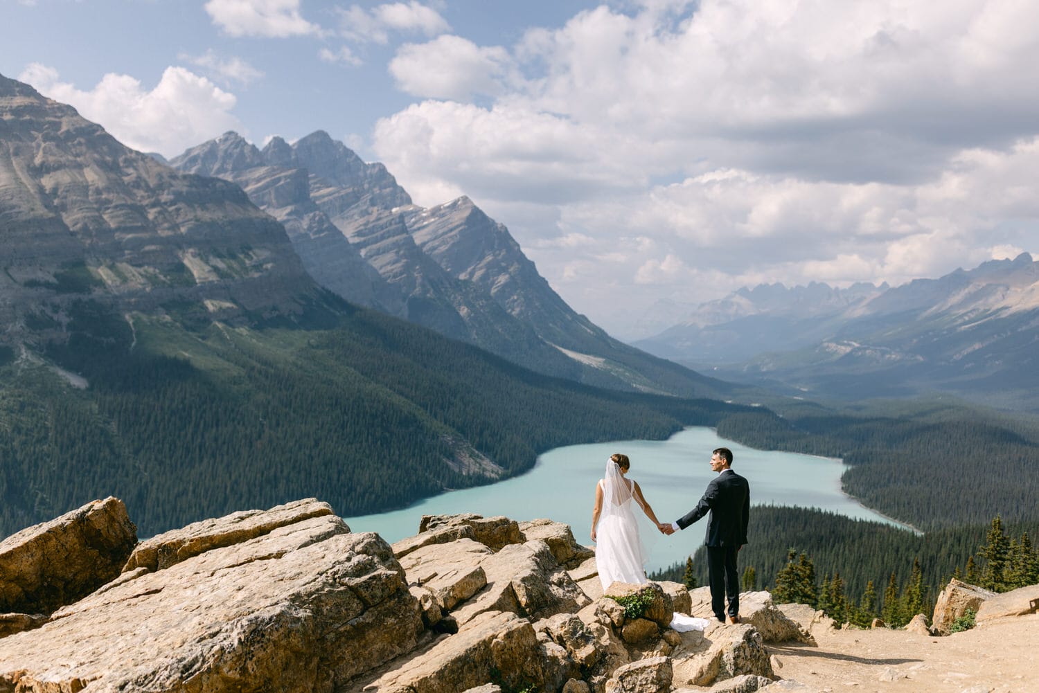 A couple stands hand in hand on a rocky outcrop with breathtaking mountain and lake views, celebrating their wedding against a stunning natural backdrop.