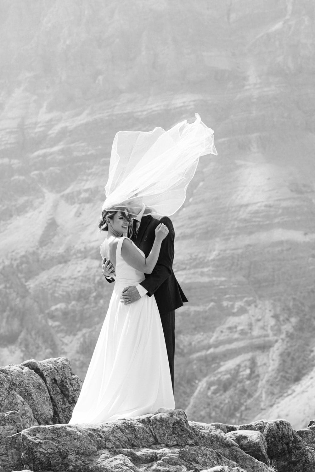 Couple embracing on rocky terrain with the bride's veil flowing in the wind, captured in elegant black and white.