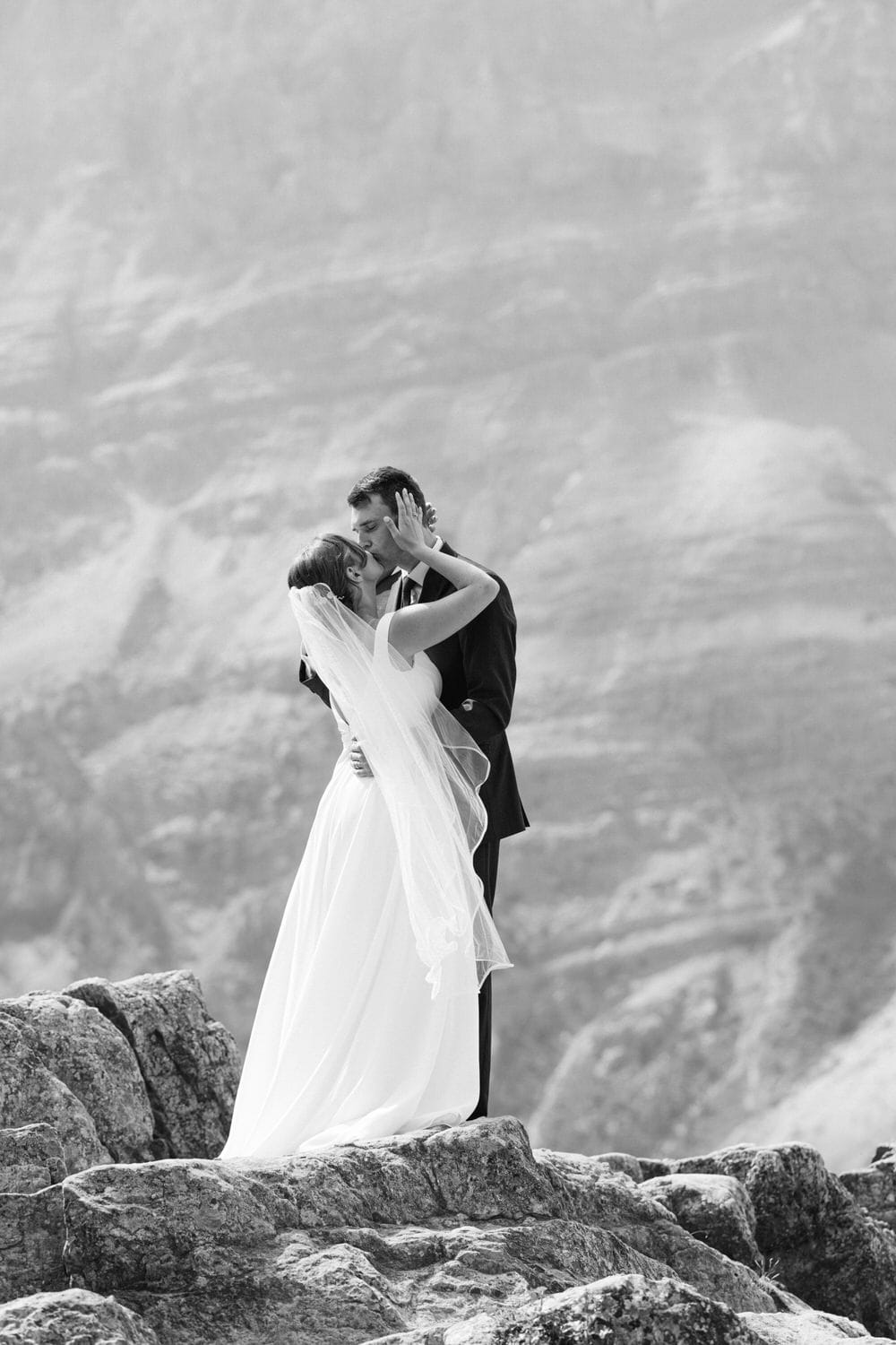 A bride and groom share an intimate moment, kissing against a stunning natural backdrop, showcasing love and connection in a black and white photograph.