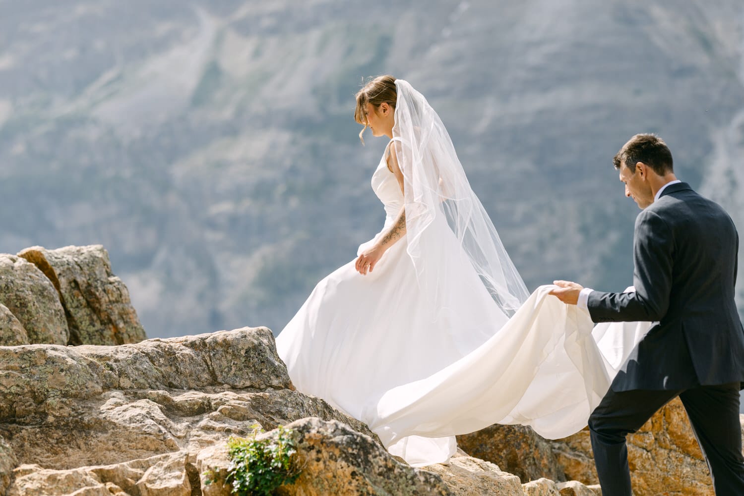 A bride gracefully walks on rocky terrain with her groom assisting her, set against a stunning mountainous backdrop.