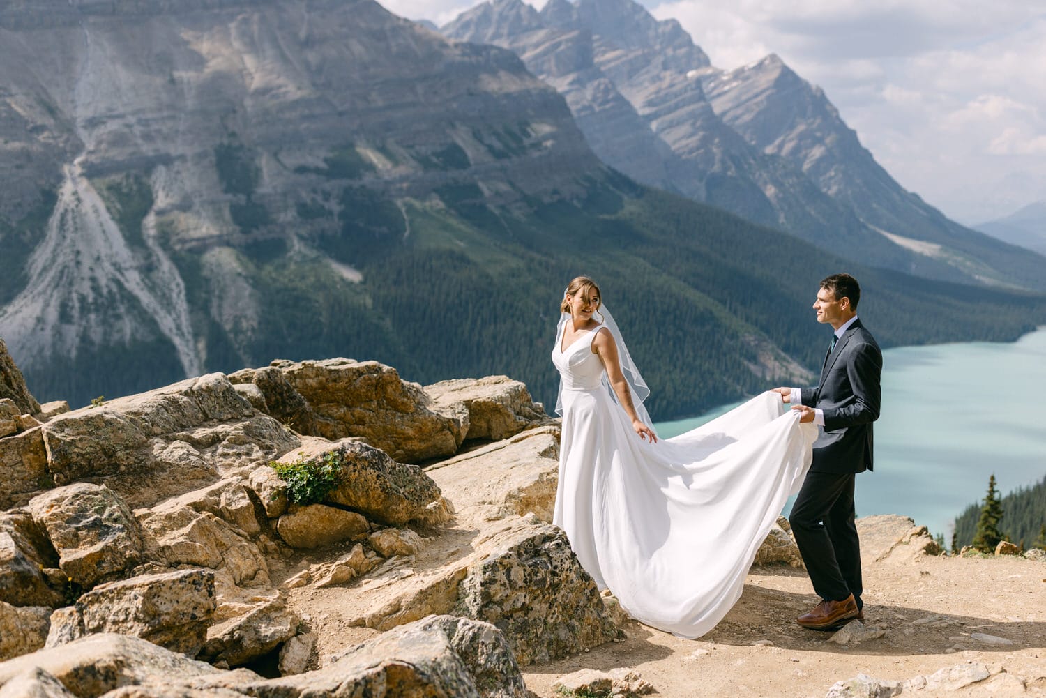 A bride in a flowing white gown and a groom hold hands on a rocky ledge surrounded by breathtaking mountains and a turquoise lake, capturing a picturesque moment of love and adventure.