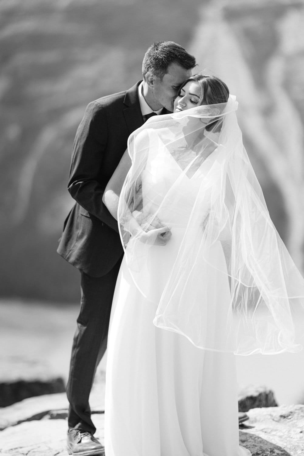 A couple sharing a loving moment, with the groom kissing the bride's forehead, as she wears a flowing veil, set against a soft blurred background.