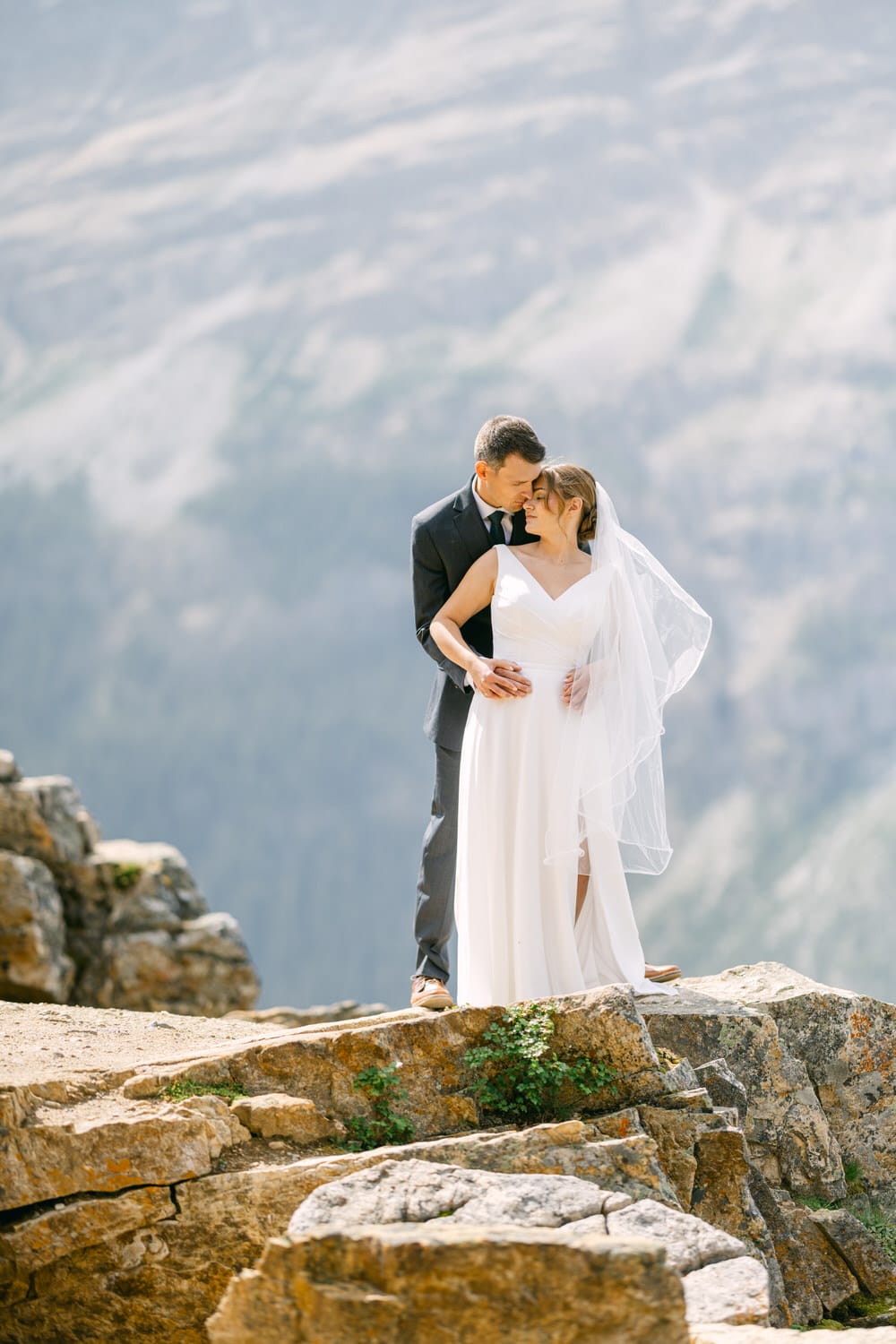 A couple embracing on a rocky ledge, with a stunning mountain backdrop, celebrating their love during a wedding ceremony.