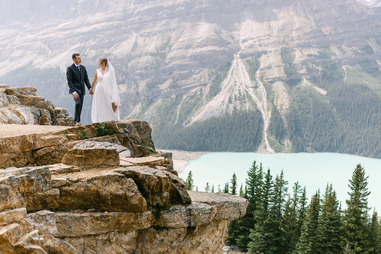 A bride and groom hold hands on a rocky ledge overlooking a serene lake and mountains, capturing a romantic outdoor wedding moment.