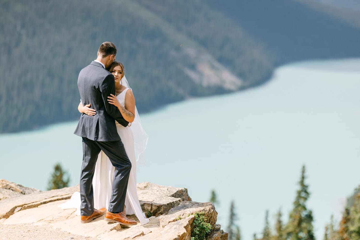 A couple embraces on a rocky ledge overlooking a serene lake and forested mountains.