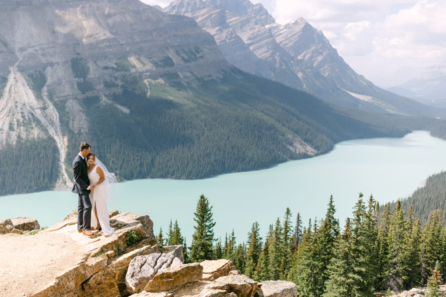 A couple embraces on a rocky ledge with stunning mountain and lake scenery in the background.