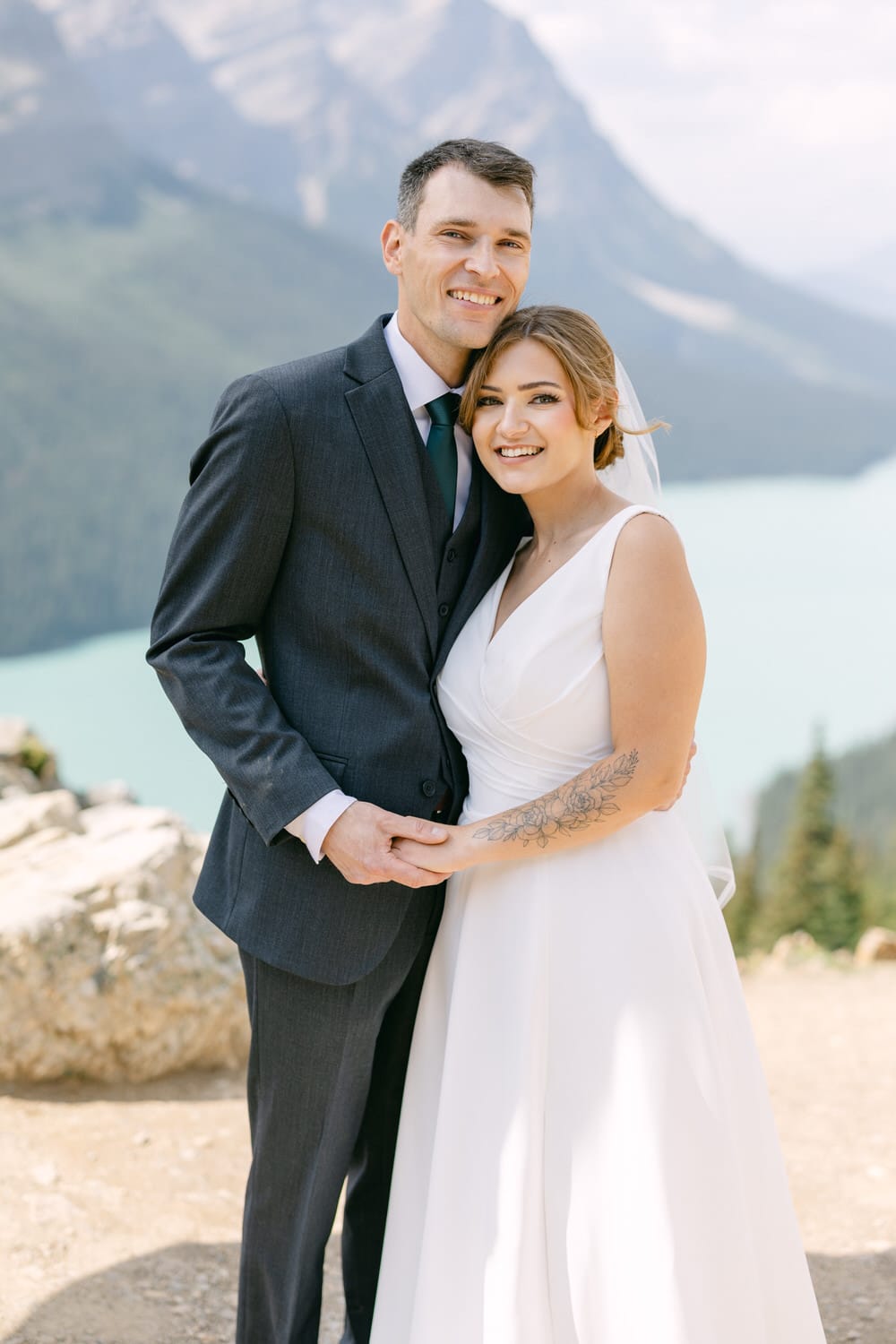 A happy couple poses together in a scenic outdoor setting, surrounded by mountains and a serene lake, showcasing love and joy on their special day.