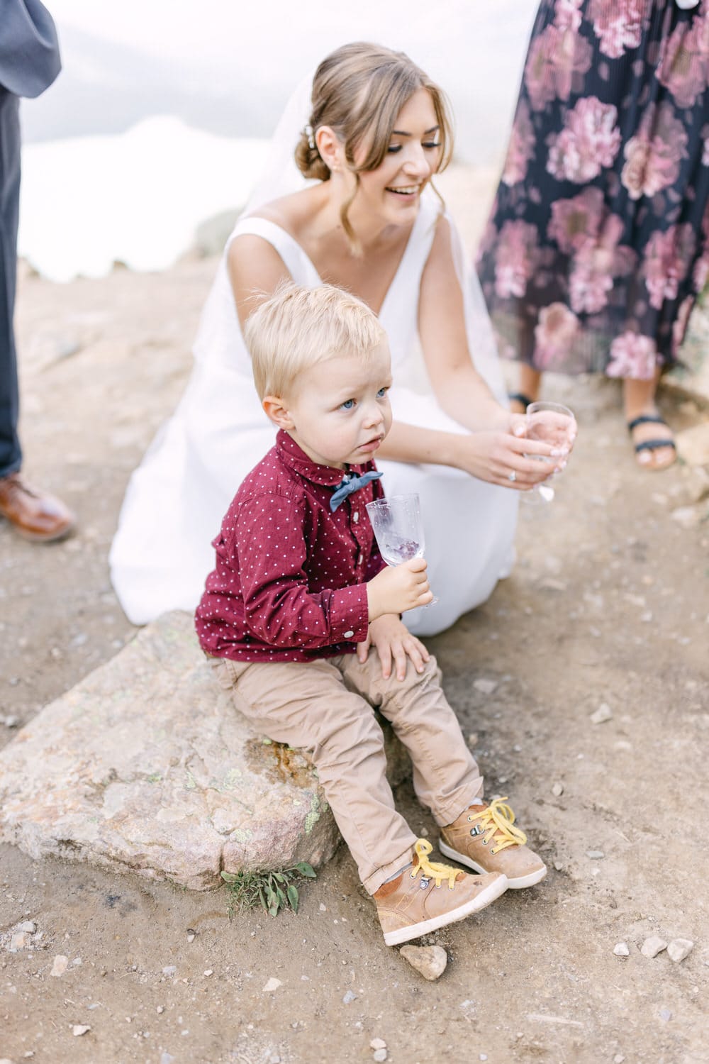 A young boy sitting on a rock, holding a glass, while a smiling bride enjoys a moment nearby.