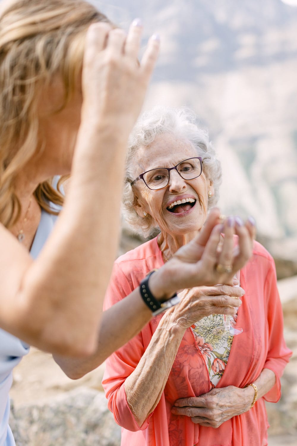 A happy elderly woman in a pink cardigan smiles while engaging with a younger woman, set against a scenic natural background.