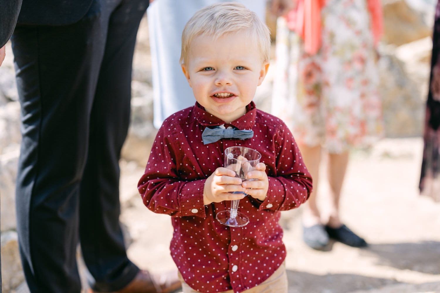 A smiling young boy in a maroon polka-dot shirt and gray bow tie holds a drink, surrounded by guests at a lively outdoor event.