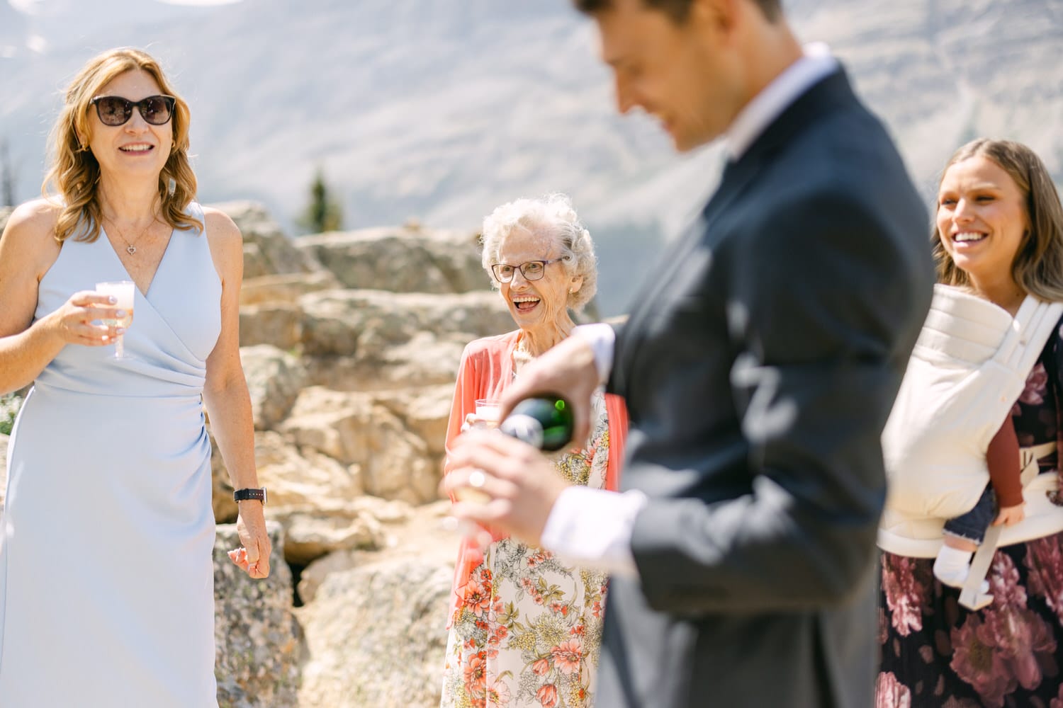 A joyous gathering featuring a woman in a blue dress, an elderly woman laughing, and a man pouring a drink, set against a mountainous backdrop.