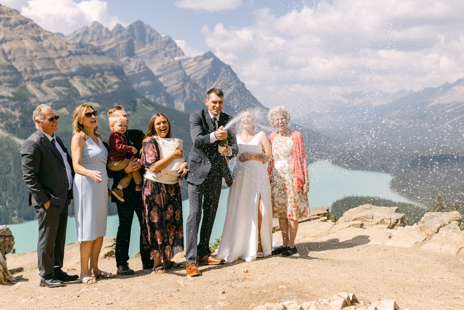 A joyful group celebrates a wedding with champagne in a scenic mountain setting, featuring a bride and groom surrounded by family against a backdrop of peaks and a glistening lake.